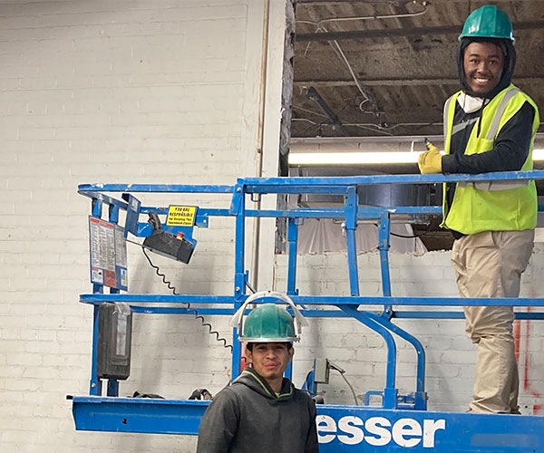 Man wearing a yellow safety vest and green hard hat while holding a thumbs up on a blue hydraulic lift as another man wearing a green hard hat stand below on the ground