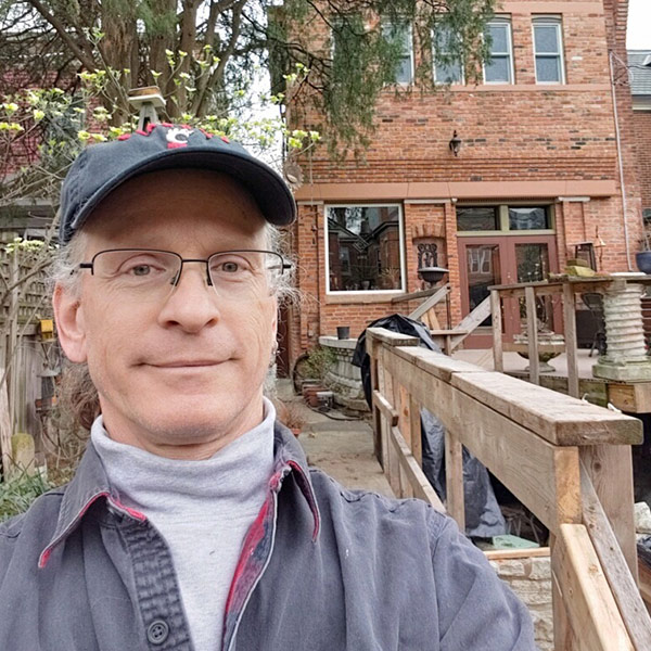 A man wearing a grey turtleneck, dark grey flannel, and a Cincinnati Bearcats hat standing by a construction area near a brick house