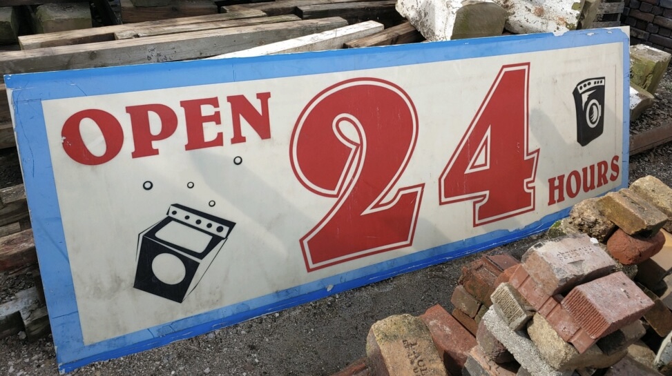 An antique “Open 24 Hours” sign with red lettering and washing machine icons, rescued by Building Value for reuse.